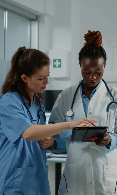 Duas médicas em scrubs azuis analisando um tablet, demonstrando colaboração e tecnologia na área da saúde.