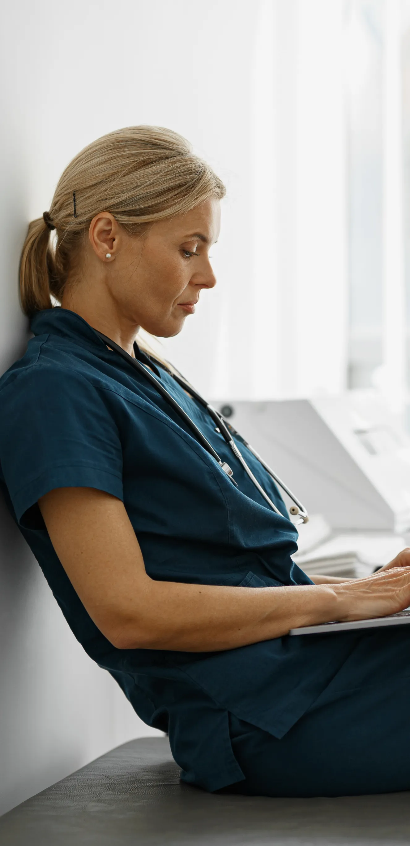 Mulher em uniforme médico sentada em um banco, usando um laptop, com expressão concentrada.